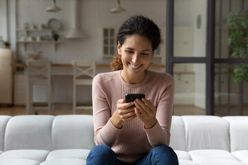 Using application. Smiling latin female spend free time sit on sofa look at smartphone screen chat...