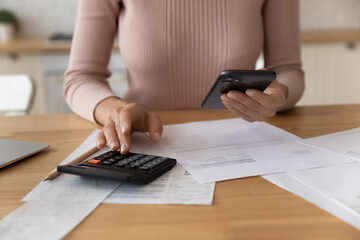 Close up of young female use phone calculator to manage financial documents bills invoices pay...