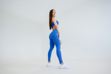 Fototapeta na wymiar Sexy young girl performs exercises with a weight in a blue tracksuit on a white background. Fitness, healthy lifestyle