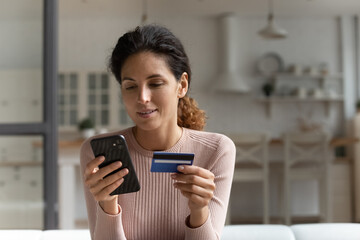 Focused concentrated millennial latina female using mobile telephone to make easy safe payment...