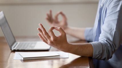 Crop close up of Caucasian man sit at desk work on laptop meditate at workplace with mudra hands....