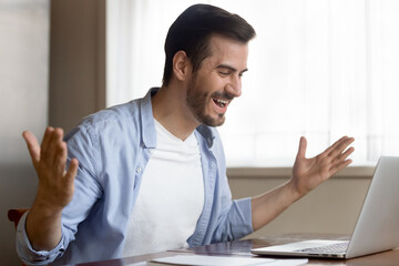 Overjoyed young Caucasian man sit at table at home office look at laptop screen triumph win lottery online. Happy millennial male feel euphoric excited read good unexpected news on computer.