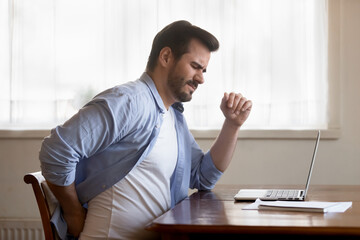 Unhappy Caucasian man sit at desk in home office work on laptop suffer from lower back muscle...