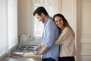 Happy young Hispanic woman hug cuddle husband cooking healthy breakfast or lunch at home kitchen. Smiling millennial wife embrace man lover preparing food, show affection love. Rent concept.
