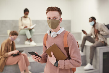 Waist up portrait of modern male student wearing mask and holding backpack while posing in school lounge looking at camera, copy space