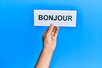 Hand of hispanic man holding french greeting bonjour word paper over isolated blue background.