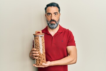 Fototapeta premium Middle age man with beard and grey hair holding jar with macaroni pasta in shock face, looking skeptical and sarcastic, surprised with open mouth