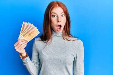 Young red head girl holding 500 norwegian krone banknotes scared and amazed with open mouth for...