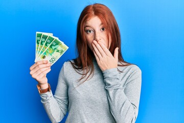 Young red head girl holding 50 israel shekels banknotes covering mouth with hand, shocked and afraid for mistake. surprised expression