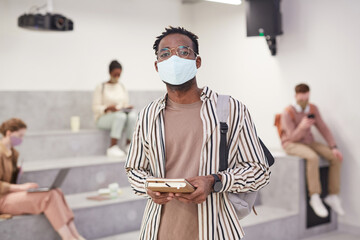 Waist up portrait of adult African-American student wearing mask and looking at camera while standing in modern school lounge, copy space