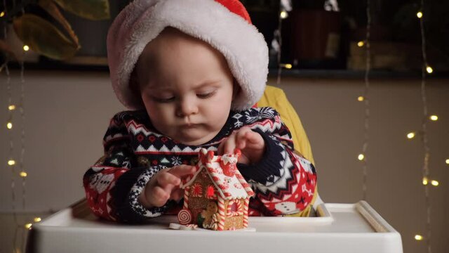 Little baby boy with sulk serious face wearing Santa hat playing with toy gingerbread house sitting in baby high chair at home. Funny baby, Christmas celebration concept.