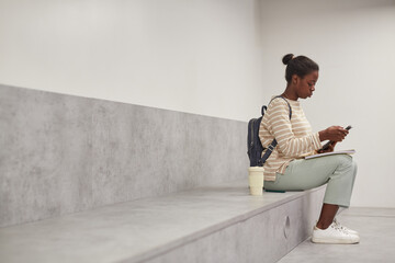 Graphic side view portrait of female African-American student using smartphone while relaxing in modern school lounge, copy space