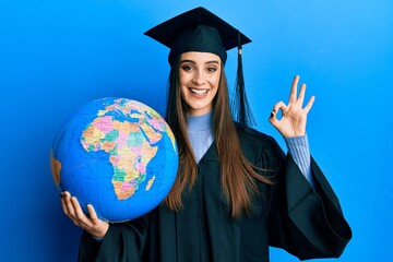 Beautiful brunette young woman wearing graduation robe holding world ball doing ok sign with fingers, smiling friendly gesturing excellent symbol