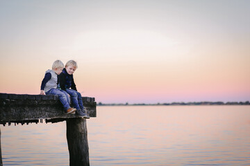 Two blonde little boys sitting together on jetty with pink sky at sunset