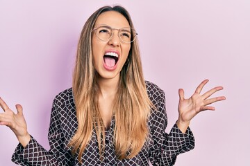 Beautiful hispanic woman wearing casual clothes and glasses crazy and mad shouting and yelling with aggressive expression and arms raised. frustration concept.