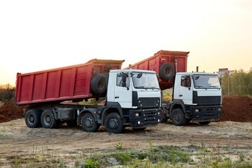 Two red dump trucks simultaneously lifted the bodies to unload the sand. Cargo transportation services. Large multi-ton truck. Unloading cargo. Construction site and machinery. Banner. Common view