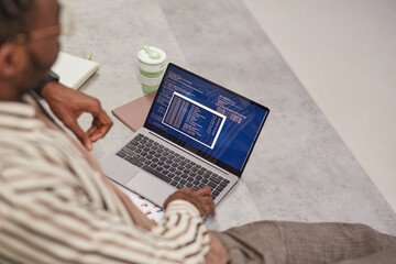 High angle view at young African-American man using laptop with IT code on screen while studying in college or working freelance, copy space