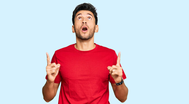 Young handsome man wearing casual red tshirt amazed and surprised looking up and pointing with fingers and raised arms.
