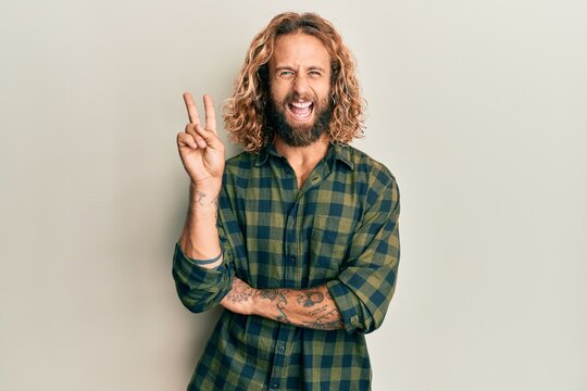 Handsome man with beard and long hair wearing casual clothes smiling with happy face winking at the camera doing victory sign. number two.