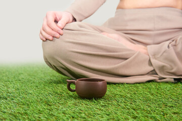 small clay tea cup on a grass carpet next to a woman sitting in lotus position on a foggy background