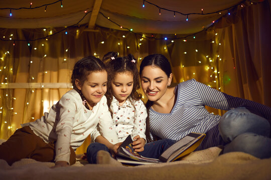 Happy family reading book together in evening. Loving mother watching picture in fairytale with two little preschool daughters children spending time in home tent with flashlights before bedding