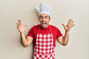 Young hispanic man wearing baker uniform celebrating mad and crazy for success with arms raised and closed eyes screaming excited. winner concept