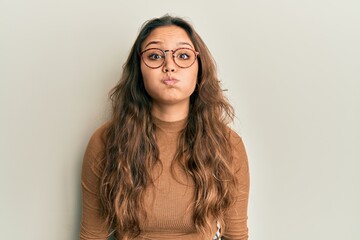 Young hispanic girl wearing casual clothes and glasses puffing cheeks with funny face. mouth...