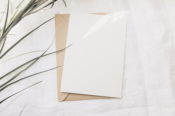 Modern summer stationery still life. Dry palm leaf on white linen table cloth. Blank greeting card mock up scene with craft envelope in sunlight. Flat lay, top view. Tropical design. Sun flare leaks.