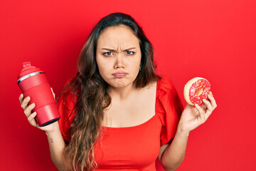 Young hispanic girl eating doughnut and drinking coffee skeptic and nervous, frowning upset because...