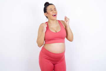 young beautiful pregnant woman in sports clothes against white wall rejoicing his success and victory clenching fists with joy being happy to achieve aim and goals. Positive emotions, feelings.
