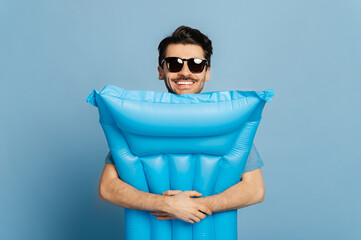 Joyful caucasian male tourist, with a sunglasses, tightly hugs a blue inflatable mattress, stands on an isolated blue background, dreams about vacation, toothy smile