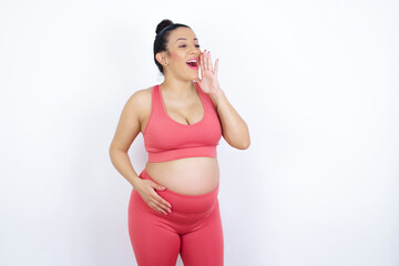 young beautiful Arab pregnant woman in sports clothes against white wall profile view, looking happy and excited, shouting and calling to copy space.