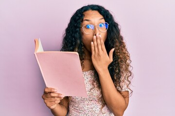 Young latin woman reading a book with glasses covering mouth with hand, shocked and afraid for...