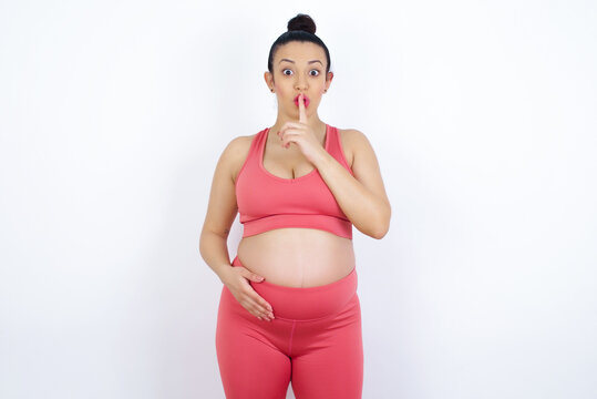 Young Beautiful Arab Pregnant Woman In Sports Clothes Against White Wall Makes Hush Gesture, Asks Be Quiet. Don't Tell My Secret Or Not Speak Too Loud, Please!