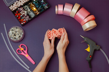 In women's hands,a hairpin with their own hands.The craft tools are set against a dark purple background. Satin pink ribbons, rhinestones, beads, scissors, glue gun, threads, glue sticks.DIY. Handmade