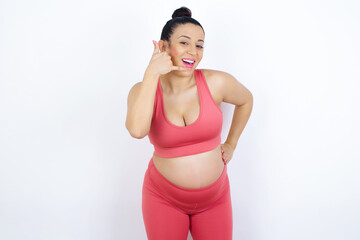 young beautiful Arab pregnant woman in sports clothes against white wall imitates telephone conversation, makes phone call gesture with hands, has confident expression. Call me!