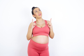 Successful friendly looking young beautiful Arab pregnant woman in sports clothes against white wall exclaiming excitedly, pointing both index fingers up, indicating something.