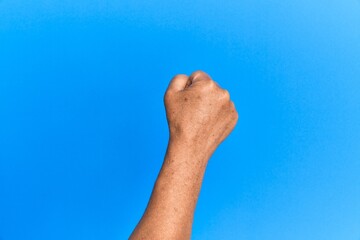 Hand of senior hispanic man over blue isolated background doing protest and revolution gesture, fist expressing force and power
