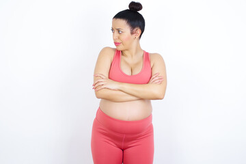 Displeased young beautiful Arab pregnant woman in sports clothes against white wall with bad attitude, arms crossed looking sideways. Negative human emotion facial expression feelings.
