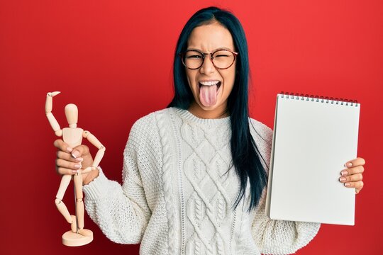 Beautiful hispanic woman holding small wooden manikin and notebook sticking tongue out happy with funny expression.