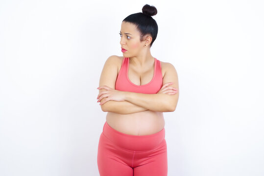 Image of upset young beautiful Arab pregnant woman in sports clothes against white wall with arms crossed. Looking with disappointed expression aside after listening to bad news.