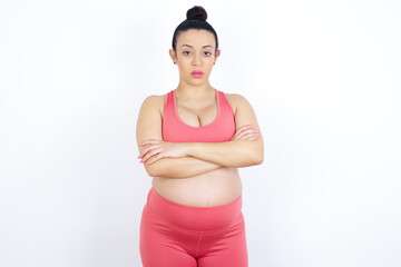 Picture of angry young beautiful Arab pregnant woman in sports clothes against white wall looking camera.