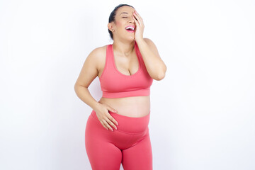 young beautiful Arab pregnant woman in sports clothes against white wall makes face palm and smiles broadly, giggles positively hears funny joke poses