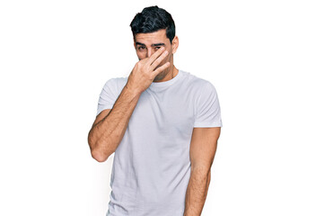 Handsome hispanic man wearing casual white t shirt smelling something stinky and disgusting, intolerable smell, holding breath with fingers on nose. bad smell