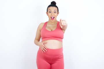 Excited positive young beautiful Arab pregnant woman in sports clothes against white wall points index finger directly at you, sees something very funny. Wow, amazing