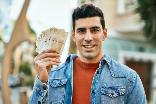 Young hispanic man smiling happy holding denmark krone banknotes at the city.