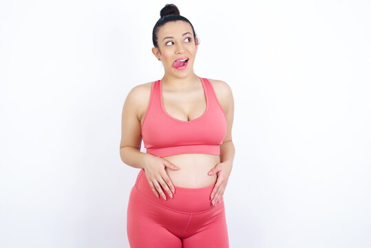 Funny young beautiful Arab pregnant woman in sports clothes against white wall makes grimace and crosses eyes plays fool has fun alone sticks out tongue.