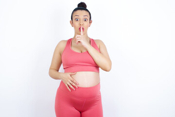 Surprised young beautiful Arab pregnant woman in sports clothes against white wall makes silence gesture, keeps finger over lips and looks mysterious at camera