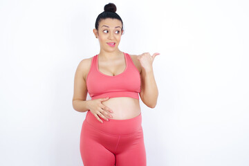 young beautiful Arab pregnant woman in sports clothes against white wall points away and gives...