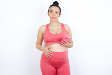 young beautiful Arab pregnant woman in sports clothes against white wall holds mobile phone uses high speed internet and social networks has online communication. Modern technologies concept